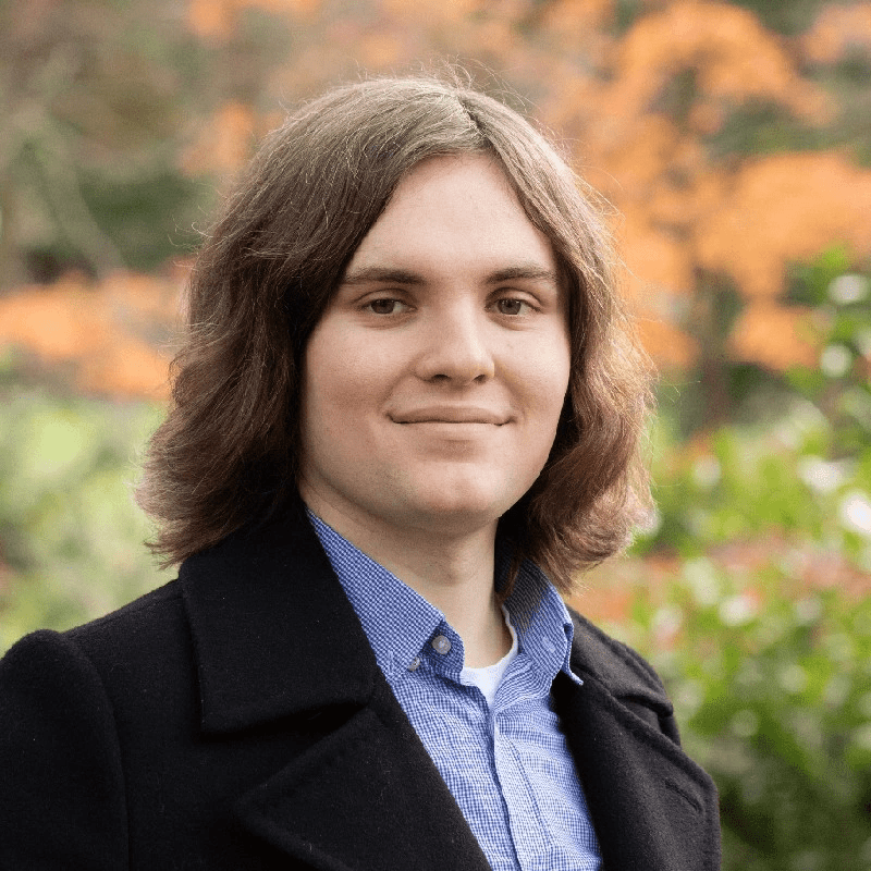 A young person with shoulder-length brown hair, wearing a blue striped shirt and a dark coat, smiles slightly whilst standing outdoors with blurred greenery and orange foliage in the background.