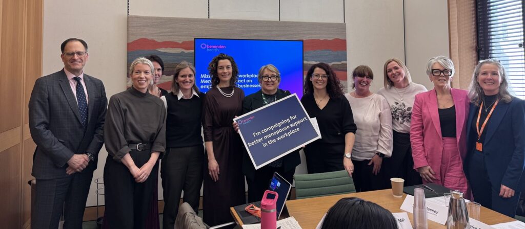 A group of ten people stand and smile in a meeting room, with a presentation screen in the background. Two people hold a sign that reads, “I’m championing for better menopause support in the workplace”.