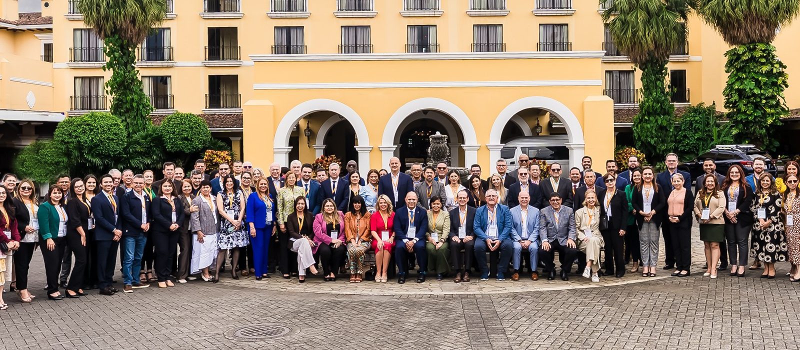 A large group of professionally dressed people is gathered for a group photo outside a yellow building with arched doorways and balconies, surrounded by greenery.