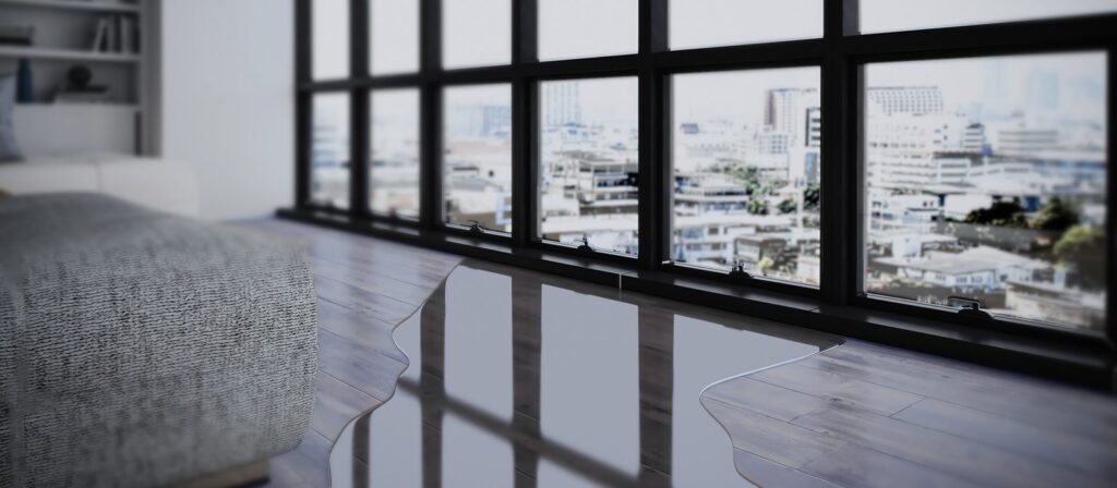 A modern living room with large windows showing a city view. There is a puddle of water on the dark wooden floor near a grey sofa, suggesting a possible leak or flooding issue.