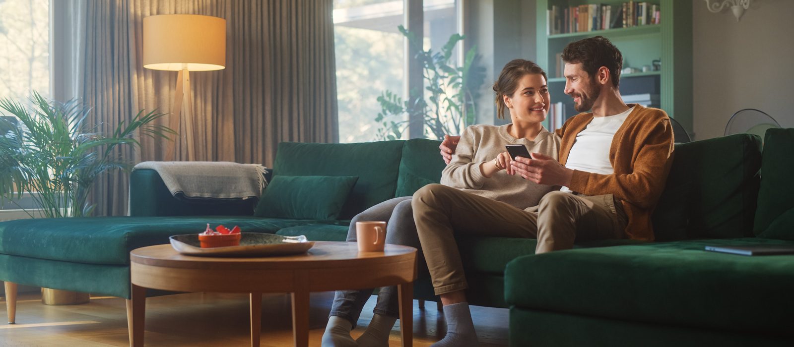 Couple Use Smartphone Device, while Sitting on a Couch in the Cozy Apartment. Boyfriend and Girlfriend Talk, do e-Shopping on Internet, Watching Funny Videos, Use Social Media, Streaming Service.