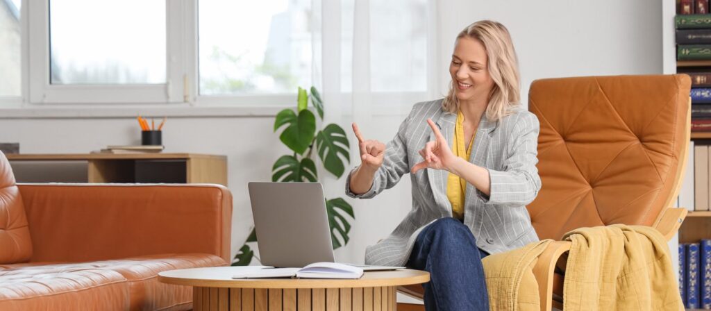 A woman sits on an armchair in a lounge, smiling and using sign language while looking at a laptop on a coffee table. An open notebook is on the table and shelves with books are in the background.