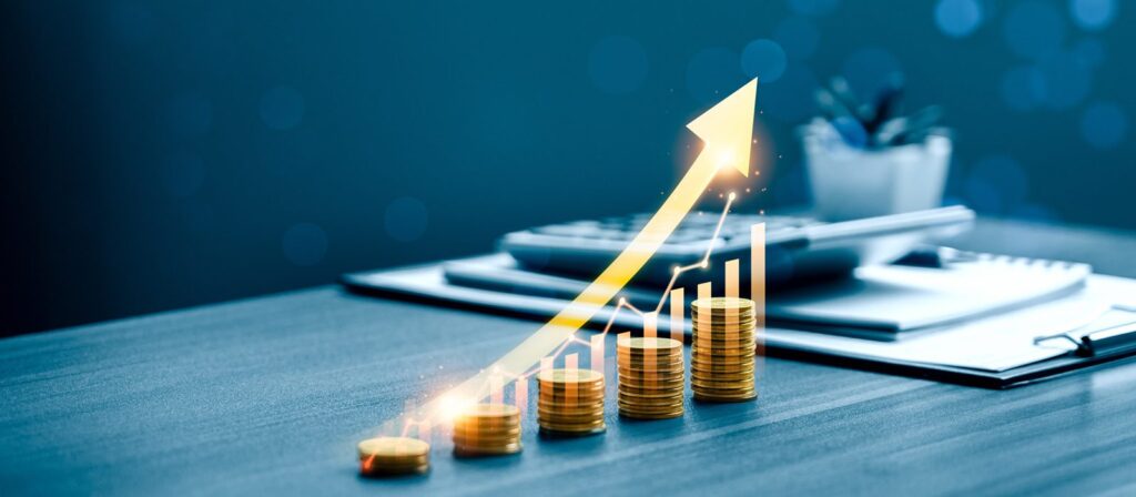 Stacks of gold coins on a table increase in height from left to right, overlaid with a glowing yellow upward arrow and bar chart, symbolising financial growth. A calculator and documents are blurred in the background.