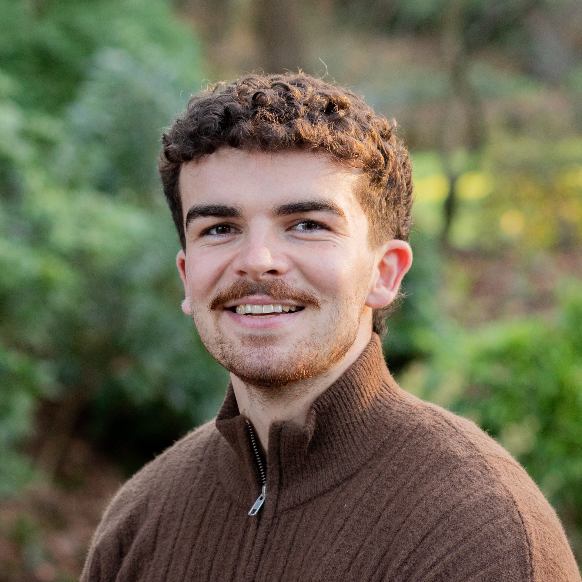 A young man with curly brown hair and a beard smiles at the camera. He is wearing a brown zip-up jumper and is outdoors, with a blurred background of greenery and trees.