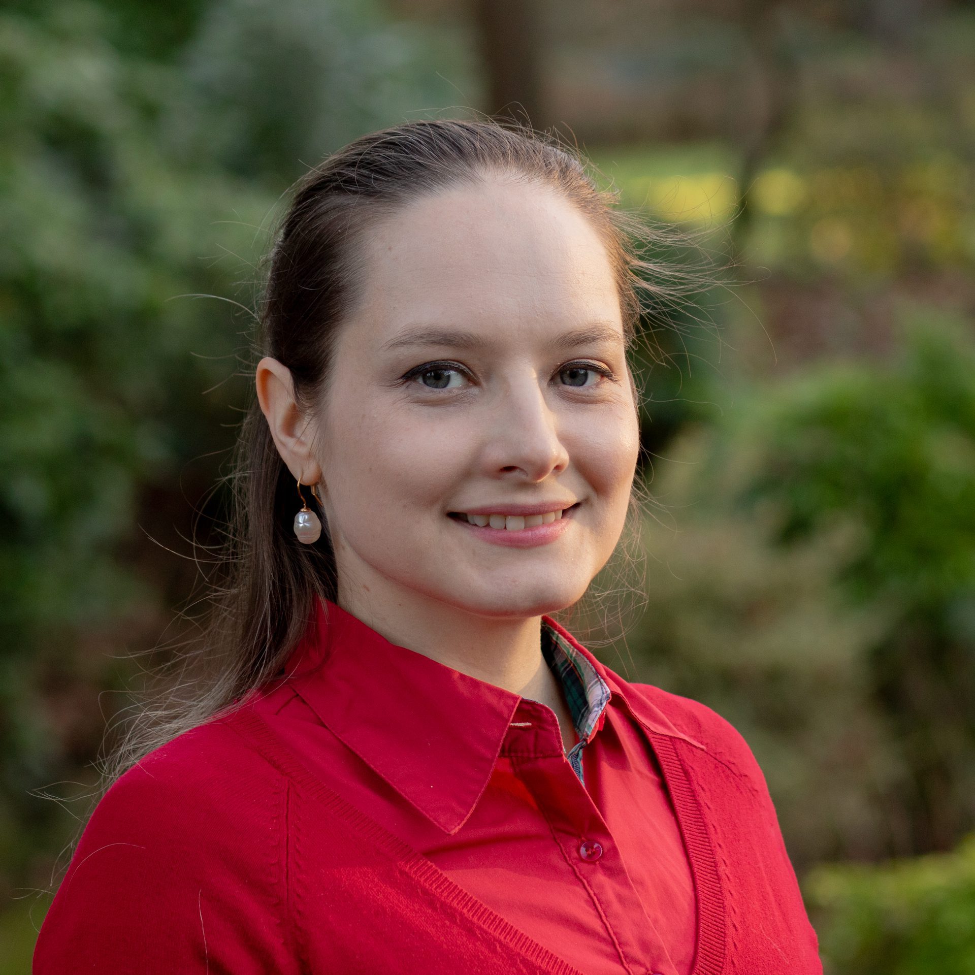 A woman with fair skin and brown hair tied back is smiling, wearing a red jumper and pearl earrings, standing outdoors with greenery in the background.