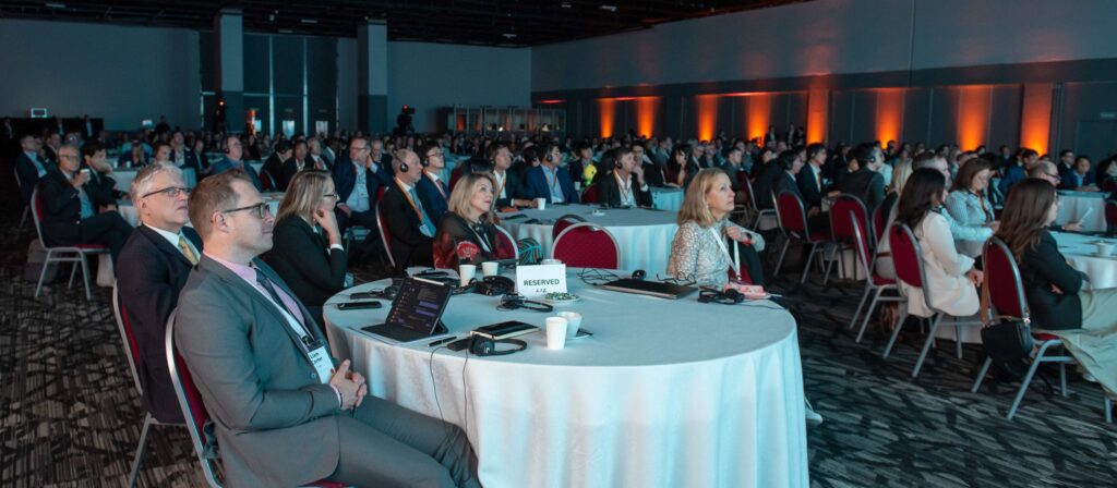A large group of people in business attire sit at round tables, attentively watching a presentation in a dimly lit conference hall with orange accent lighting.
