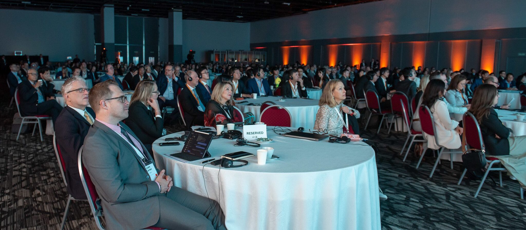 A large group of people in business attire sit at round tables, attentively watching a presentation in a dimly lit conference hall with orange accent lighting.