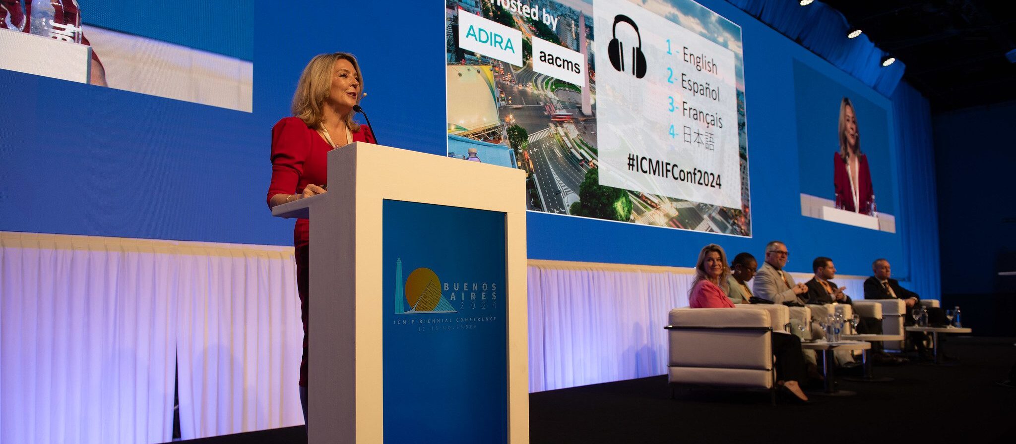 A woman speaks at a lectern on a stage with a screen behind her showing conference details. Four panellists sit on white chairs to the right. The event appears to be held in Buenos Aires, as indicated on the lectern.