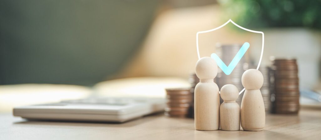 Wooden figures representing a family stand in front of stacked coins, a calculator, and a shield with a tick, symbolising financial security and protection.