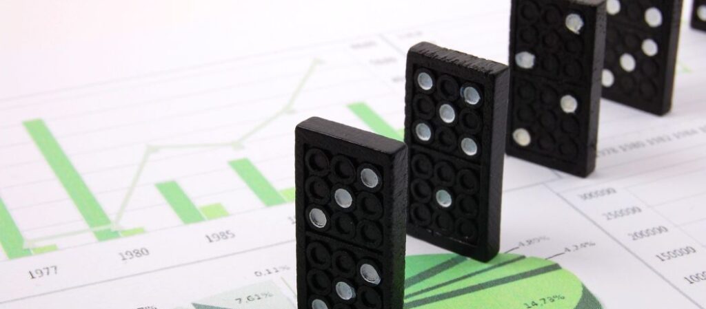 Several black dominoes stand in a row on top of financial charts with green bar and pie graphs, illustrating the concept of a domino effect in financial data or business impact.