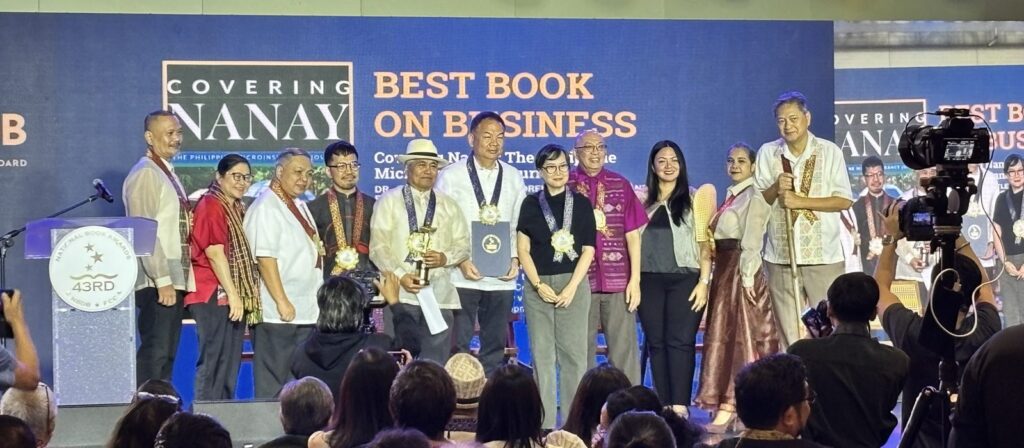 A group of people stand on stage, some holding trophies and wearing medals, during an award ceremony for “Best Book on Business.” A backdrop behind them displays the event title and logo as photographers capture the moment.