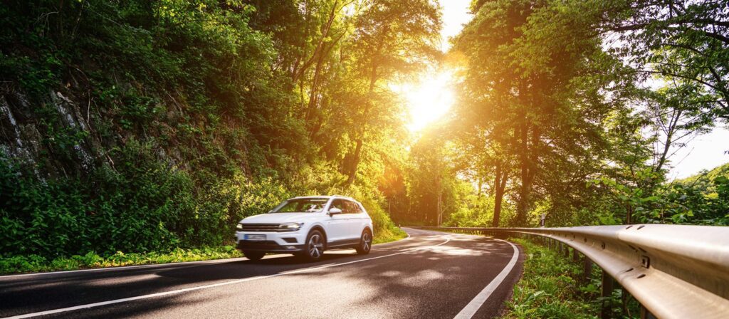 A white SUV drives along a winding road surrounded by lush green trees, with sunlight streaming through the foliage on a bright day.