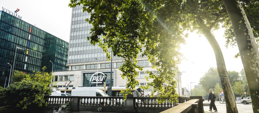 Sunlight filters through leafy trees beside a modern city street, with glass and concrete office buildings, parked vans, and pedestrians visible in the background.