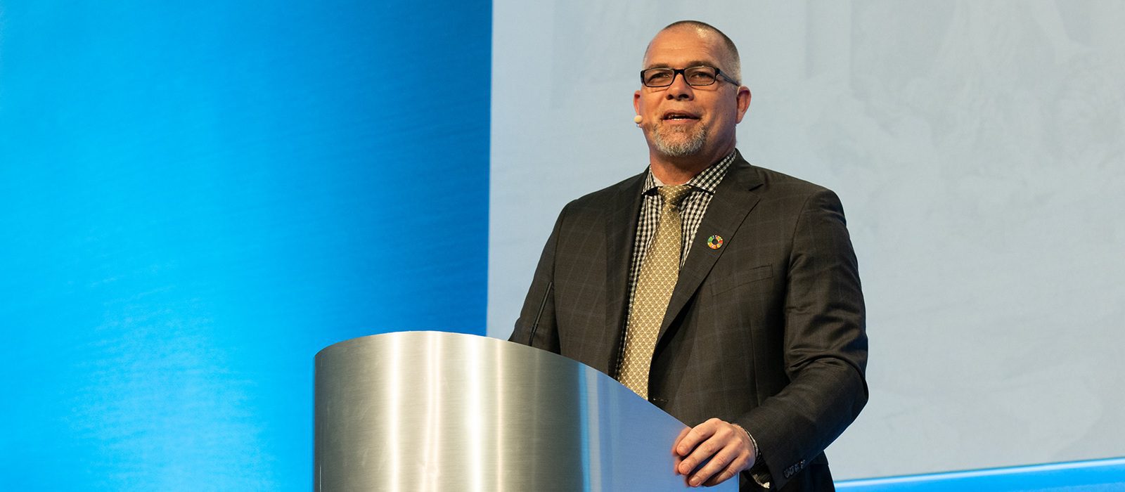 A man in a suit, tie, and glasses stands at a lectern, speaking in front of a blue and white background at what appears to be a professional conference or event.