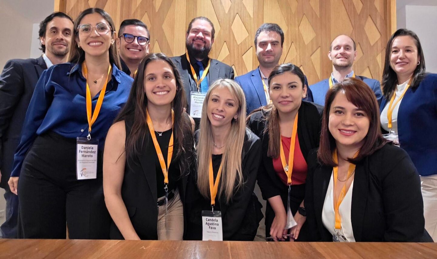 A group of eleven professionally dressed people with name badges and yellow lanyards pose together indoors in front of a wooden wall, smiling at the camera.