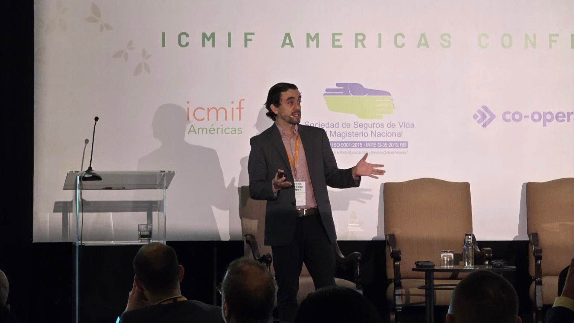 A man in a suit stands on stage giving a presentation at the ICMIF Americas Conference, gesturing with his hands. Behind him are logos and an empty lectern; some audience members are visible in the foreground.