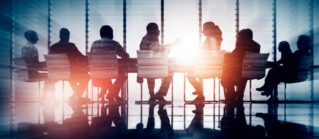 A group of people sit around a conference table in a modern office with large windows, silhouetted against bright sunlight streaming through the glass.