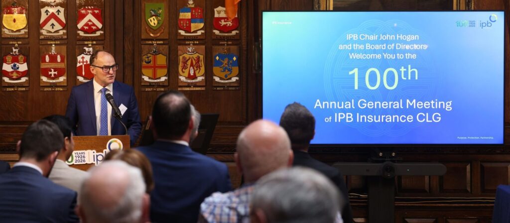 A man speaks at a lectern in front of an audience. Behind him is a wooden wall with shields and a large screen displaying, 100th Annual General Meeting of IPB Insurance CLG.