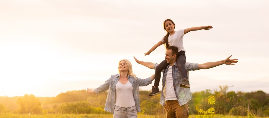 A smiling family outdoors at sunset; a woman and man stand with arms outstretched, while a young girl sits on the man's shoulders, all appearing joyful and relaxed in a grassy, sunlit field.