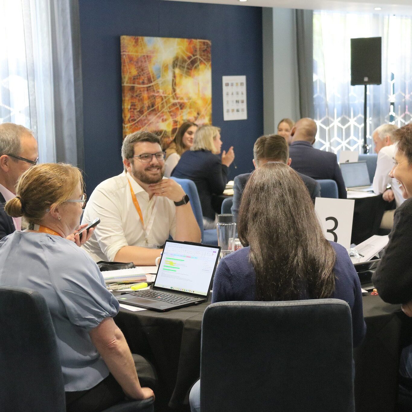 A group of people sit around a table in a modern meeting room, engaged in discussion with laptops and notes. Other groups can be seen talking at nearby tables.