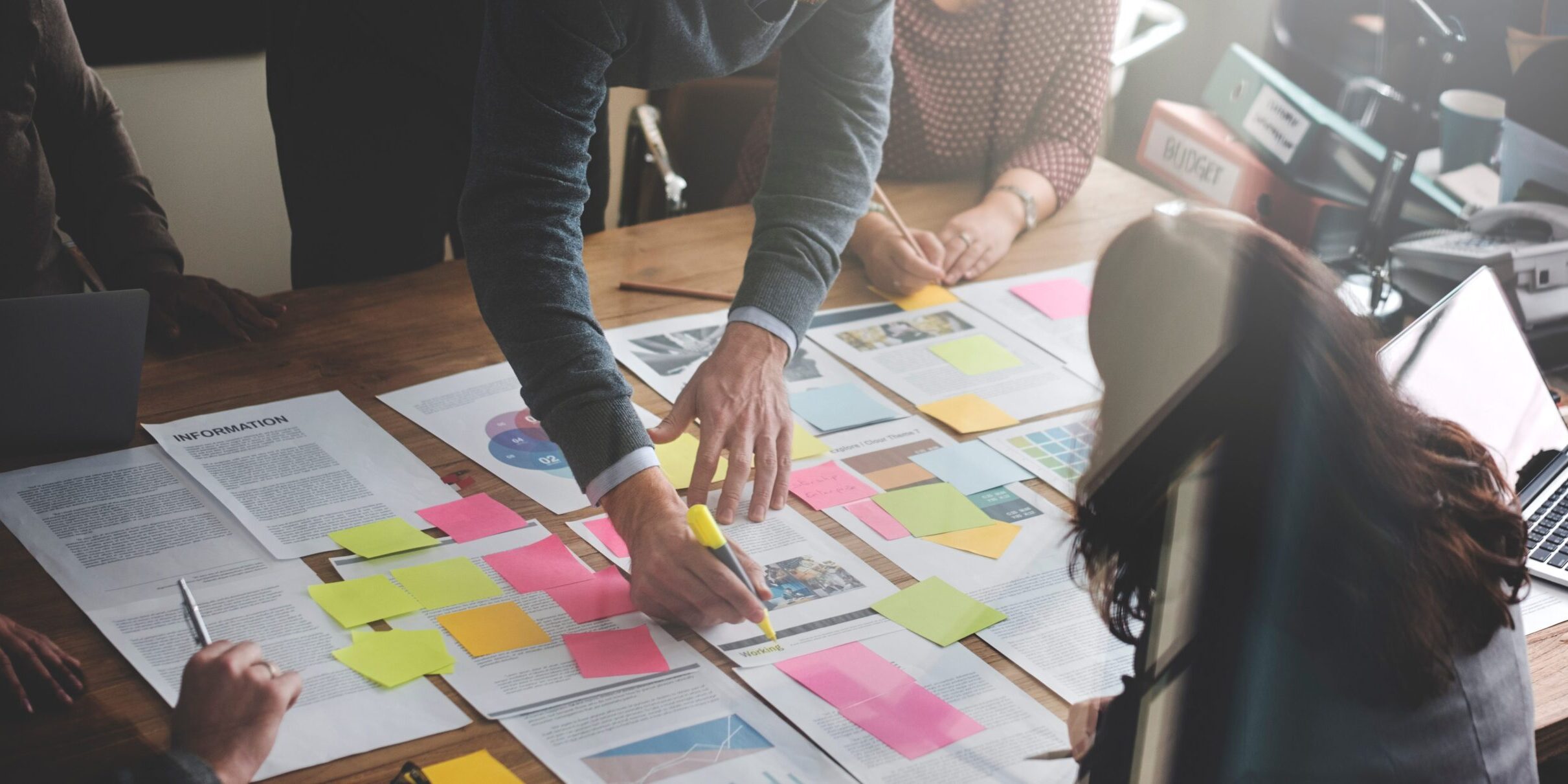 Several people collaborate at a table covered with papers and colourful sticky notes. One person is marking a document with a highlighter, while others lean in and discuss, suggesting a team brainstorming or planning session.