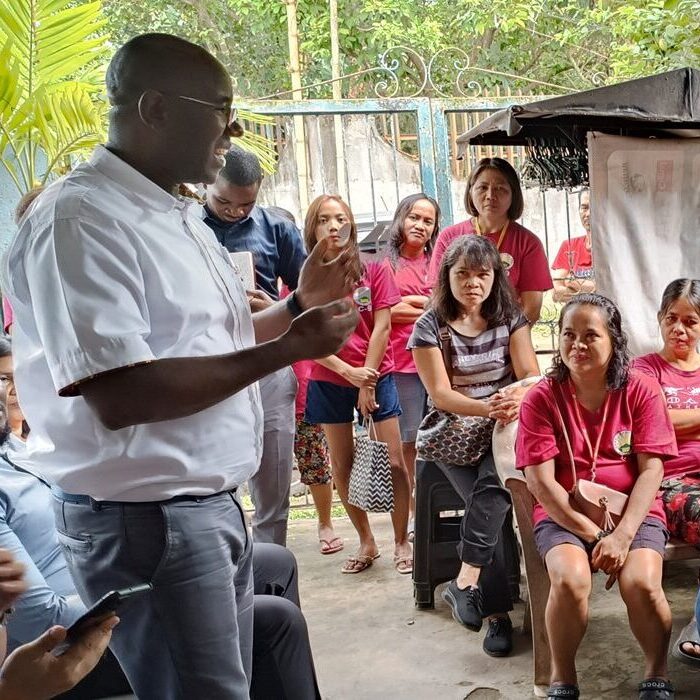 A man in a white shirt speaks to a group of attentive women, most wearing red shirts, gathered outdoors near a fence and greenery. Some are seated while others stand, listening closely.