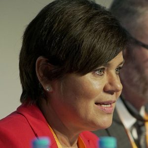 A woman with short brown hair, wearing a red blazer, sits at a table and speaks, with a blurred man in the background. Several water bottles are visible in the foreground.