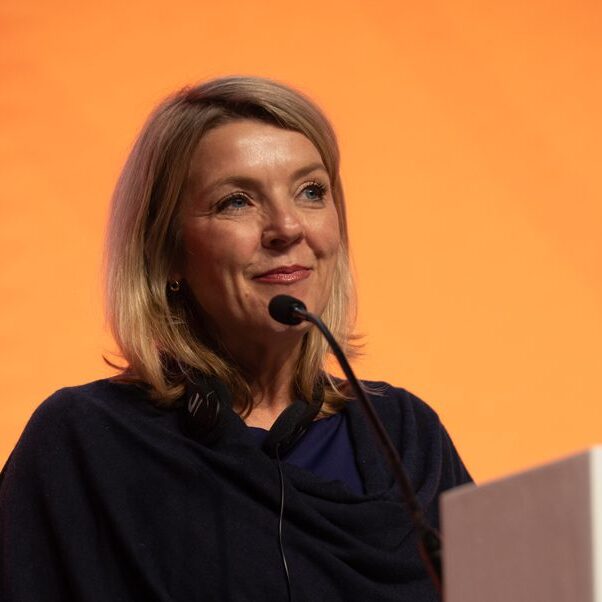 A woman with blonde hair speaks into a microphone at a podium, smiling slightly. She wears a dark top and the background is a solid orange color.