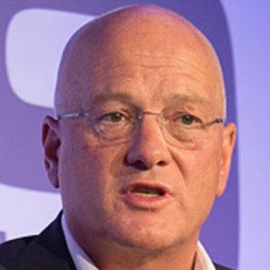 A middle-aged, bald man wearing glasses and a suit jacket speaks in front of a blue and white background.