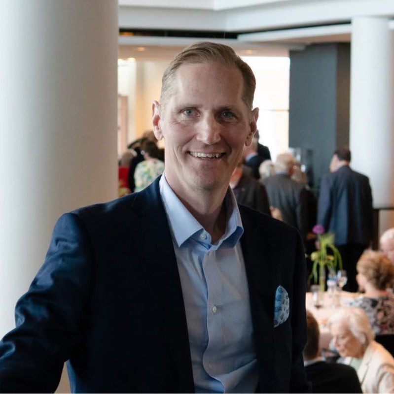 A man in a navy suit and light blue shirt smiles at the camera in a bright, modern room with large windows and people socialising in the background.