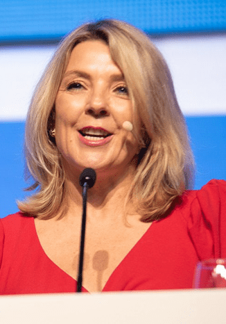 A woman with shoulder-length blonde hair, wearing a red top and a headset microphone, speaks at a lectern with a blue background. A water bottle and glass are visible in front of her.