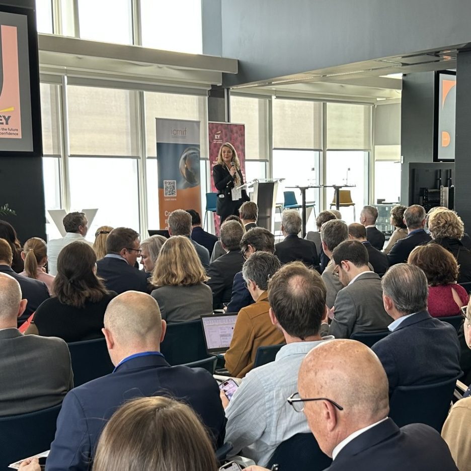 A woman stands at a lectern speaking to a seated audience in a bright, modern conference room with large windows and banners in the background.